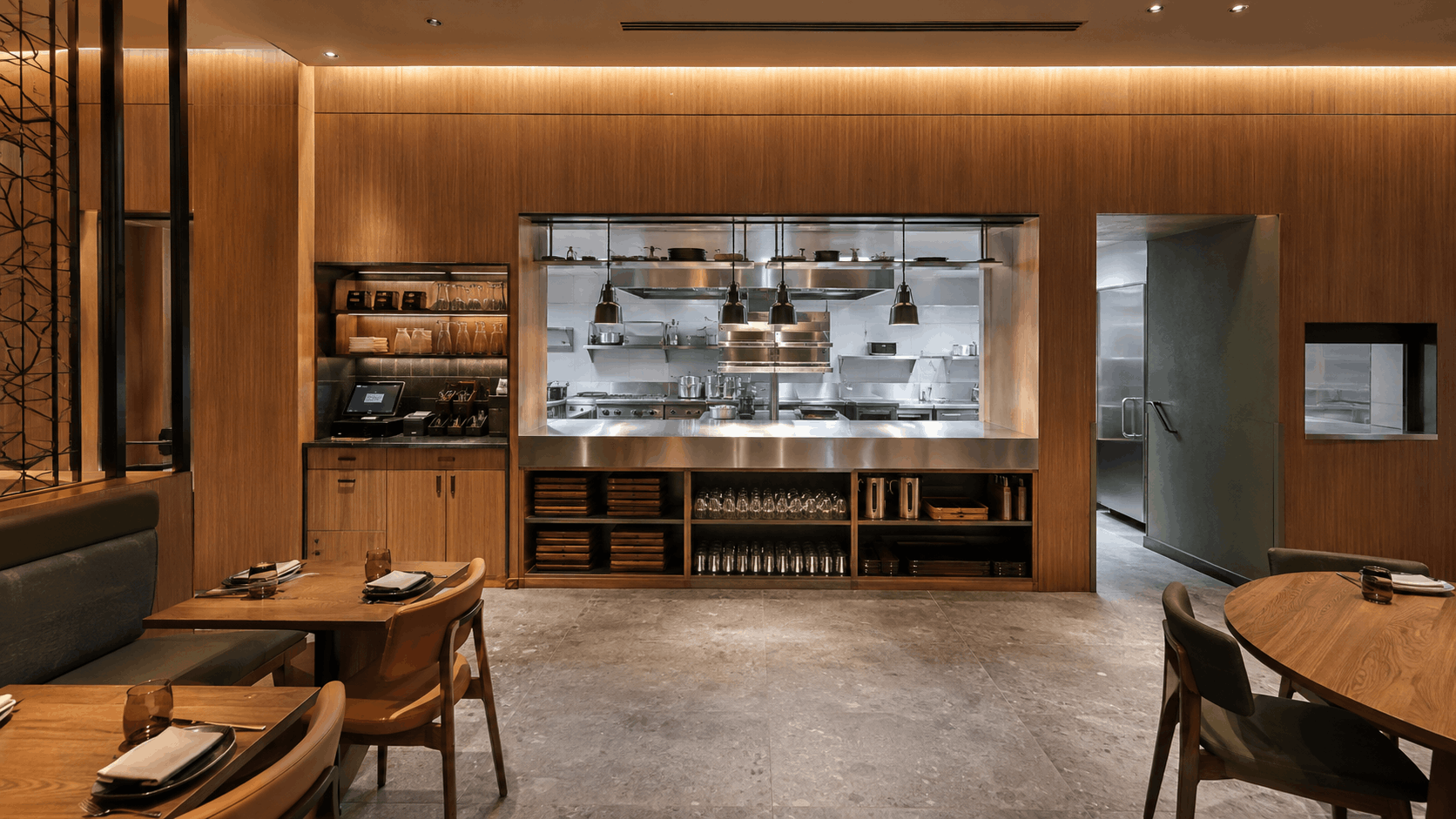 Open commercial kitchen visible from a warm, wood-panelled restaurant dining room through a stainless steel pass counter